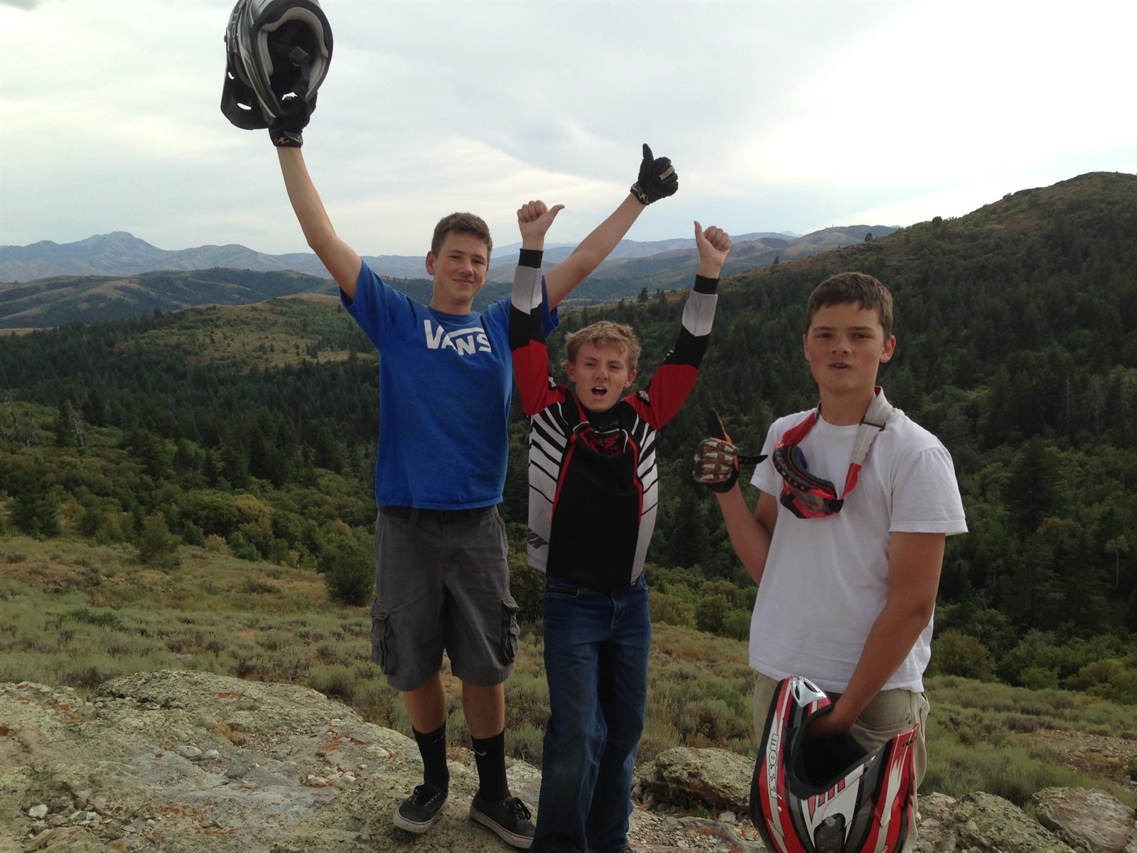 Three boys celebrating with helmets raised on a mountain overlook