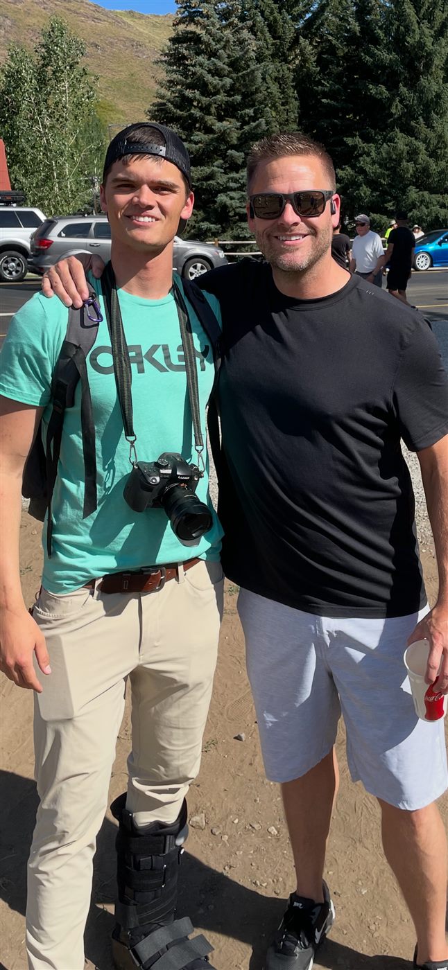 Caden with a Lumix camera around his neck standing beside a friend, wearing a walking boot
