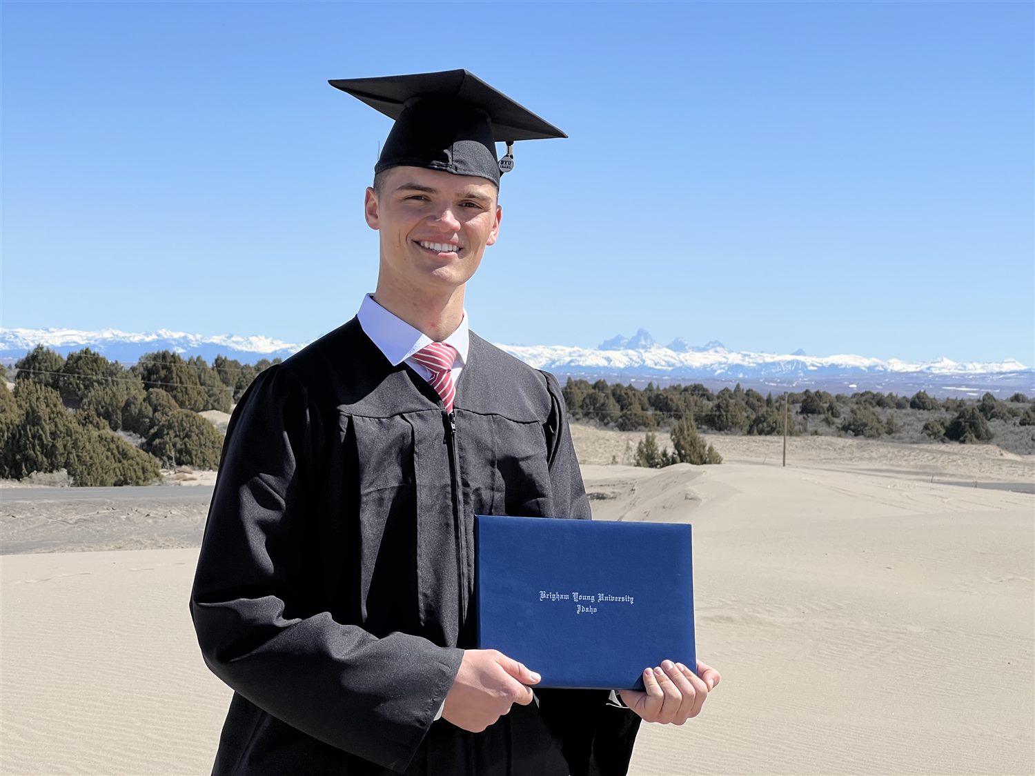 Caden in cap and gown holding his BYU-Idaho diploma on sand dunes with the Tetons behind