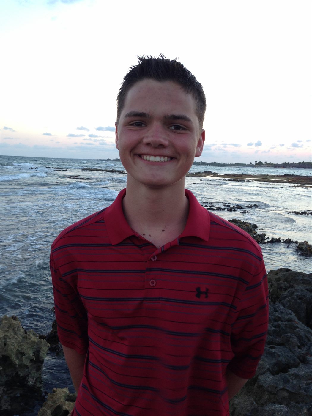 Caden in a red polo smiling at a rocky ocean shore
