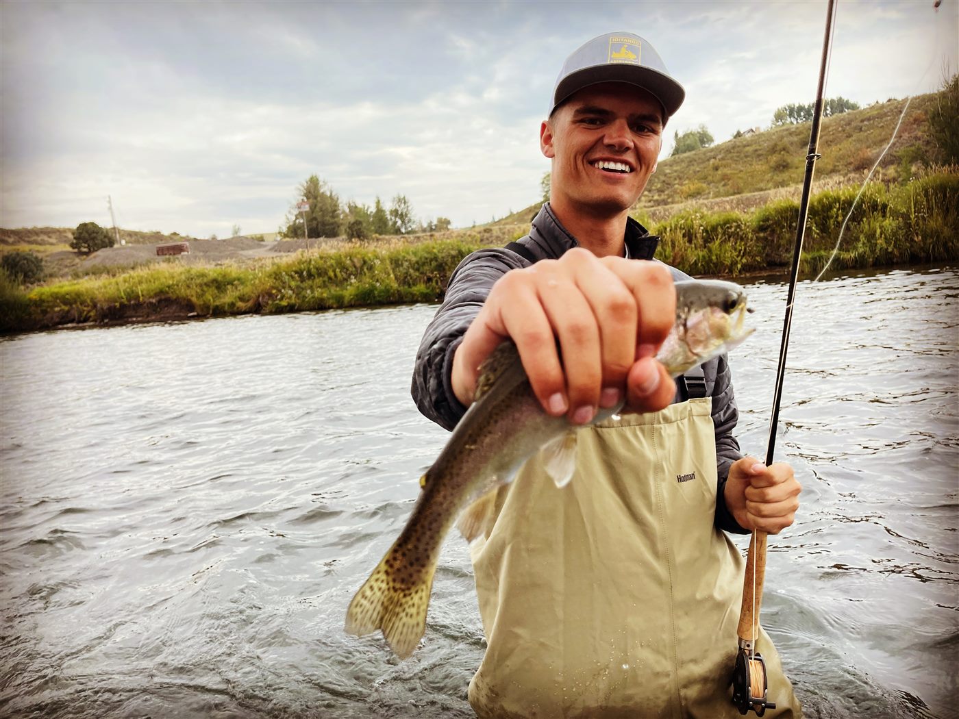 Caden in waders holding a trout toward the camera in the middle of a river